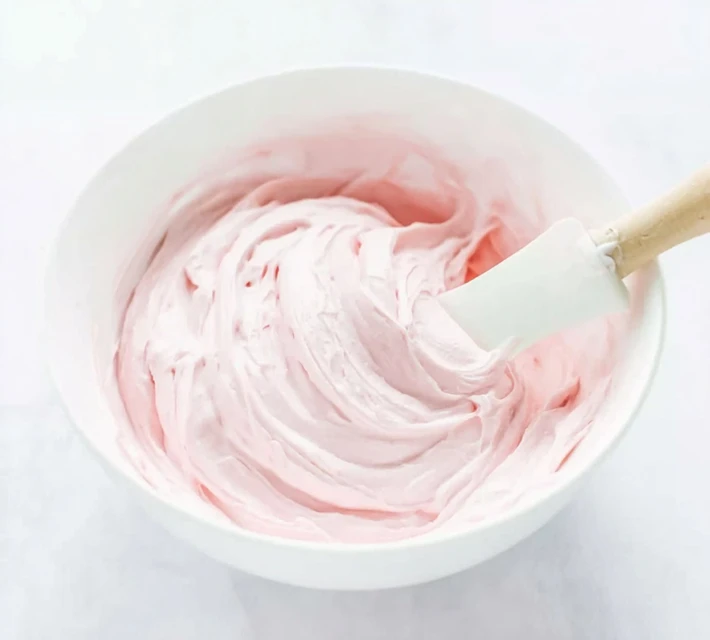 Mixing strawberry marshmallow frosting in a white bowl with a spatula