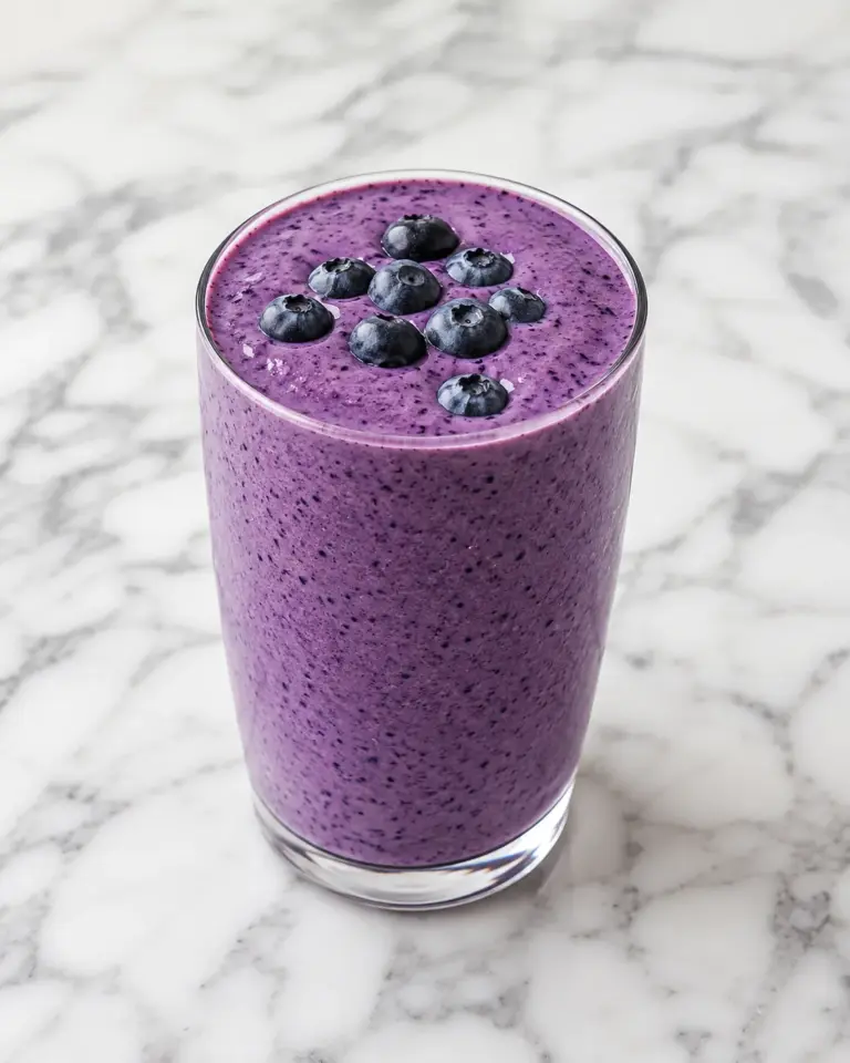 Creamy blueberry smoothie in a clear glass on a white marble surface with fresh blueberries.