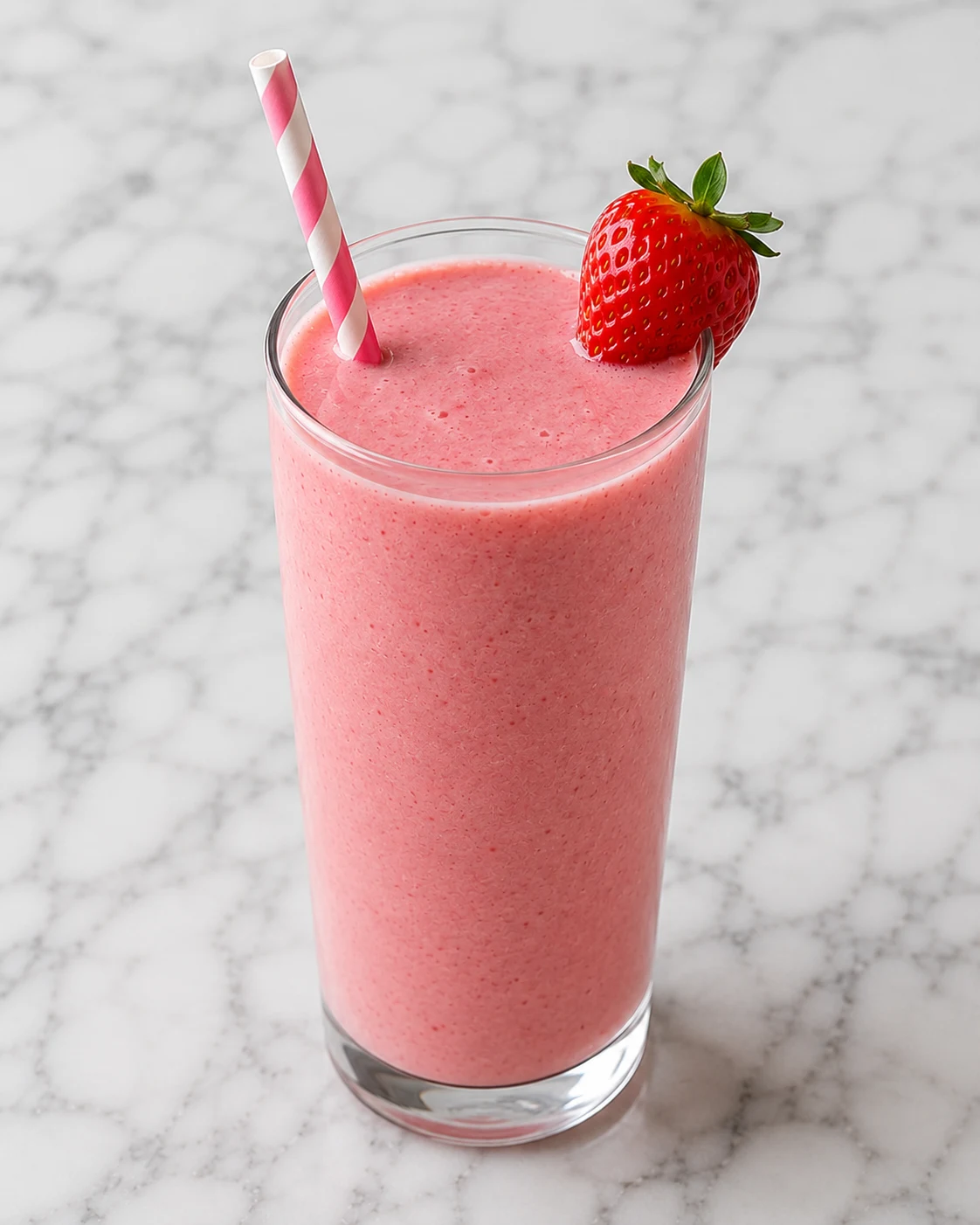 Creamy strawberry smoothie in a tall glass with a striped straw and fresh strawberry garnish on a white marble surface.
